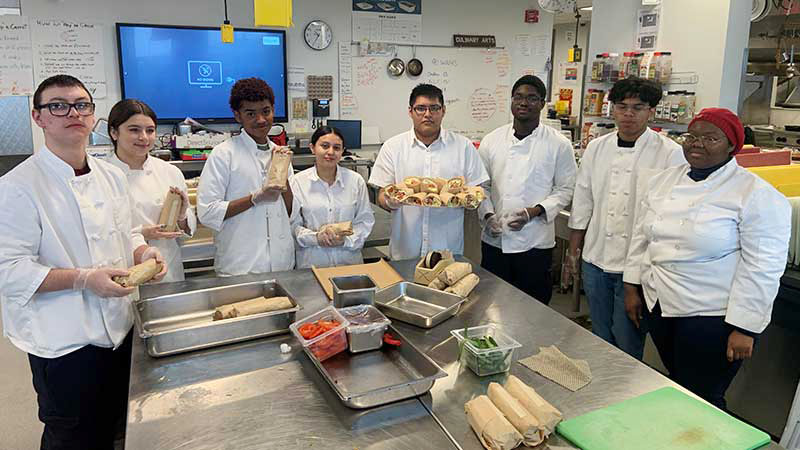 Students in uniform in the culinary arts kitchen holding gourmet sandwiches