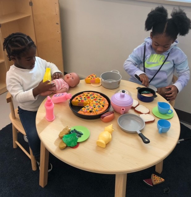two students sitting at a miniature kitchen table with dishes and pans